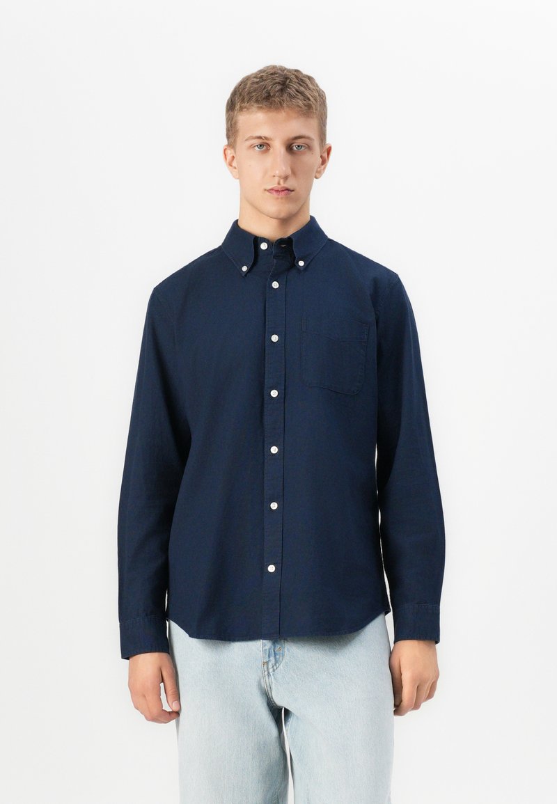 Joven vestido con una camisa de botones azul marino con un bolsillo en el pecho y jeans de color azul claro, de pie frente a un fondo blanco liso.