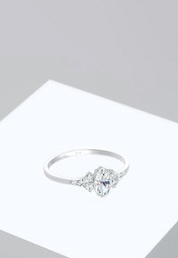 Anillo de plata con diamante ovalado en el centro flanqueado por diamantes laterales triangulares sobre una plataforma geométrica blanca sobre un fondo gris.