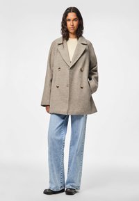 Cappotto grigio doppiopetto in misto lana, con grandi bottoni, ampi rever e vestibilità morbida, abbinato a jeans a gamba larga di colore blu chiaro.