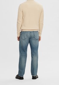 Světlý béžový pletený svetr se žebrovanou texturou, doplněný džínami středního praní s rovnými nohavicemi a zadními kapsami. Černé boty.