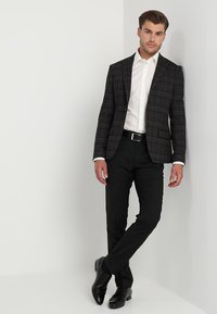Blazer a quadri in tonalità scure con rever a punta, camicia bianca, pantaloni neri e scarpe nere lucide. Texture lisce, vestibilità su misura.