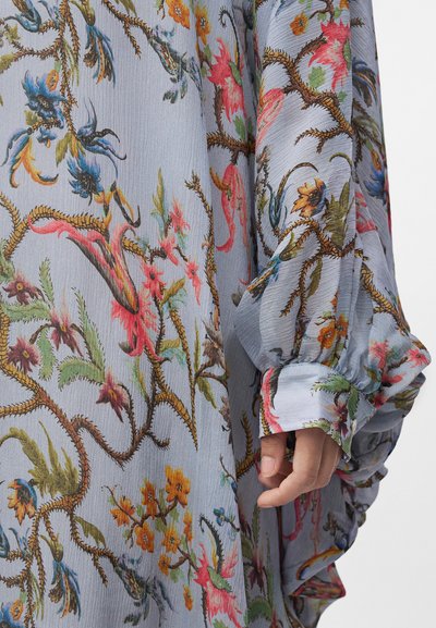 Manche avec poignet froncé sur un tissu bleu clair texturé à motifs floraux colorés et de vignes épineuses.
