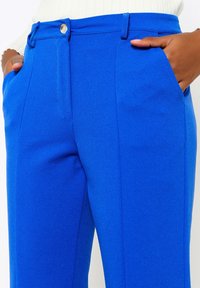 Blå byxor av mjukt tyg med platt framstycke, hällor för bälte och sidofickor. Förstärkta sömmar och en knappstängning.