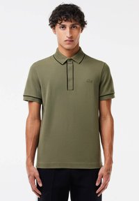 Polo de color verde oliva con mangas cortas, tapeta de tres botones y logotipo bordado. Presenta detalles en negro contrastantes en el cuello y los puños.