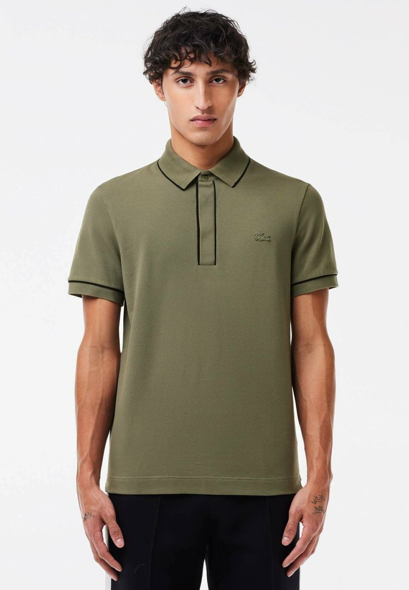 Polo de color verde oliva con mangas cortas, tapeta de tres botones y logotipo bordado. Presenta detalles en negro contrastantes en el cuello y los puños.