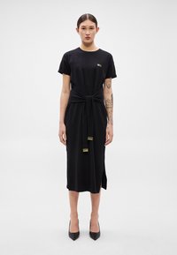 Μαύρο midi φόρεμα με κοντά μανίκια, λεπτομέρεια δέσιμου στη μέση και χρυσές ακτίνες. Ομαλό ύφασμα, ευθεία κοπή, πλευρική σκίσιμο, συνδυασμένο με μαύρα τακούν�ια.