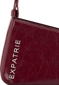 Burgundivörös, műbőr keresztpántos táska textúrázott felülettel. Az oldalon fémes "EXPATRIÉ" logó található. Háromszögletű forma, állítható pánttal.