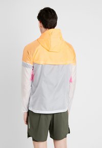 Chaqueta ligera y multicolor que presenta paneles en naranja, gris y rosa, con capucha y tejido texturizado. Incluye detalles reflectantes.