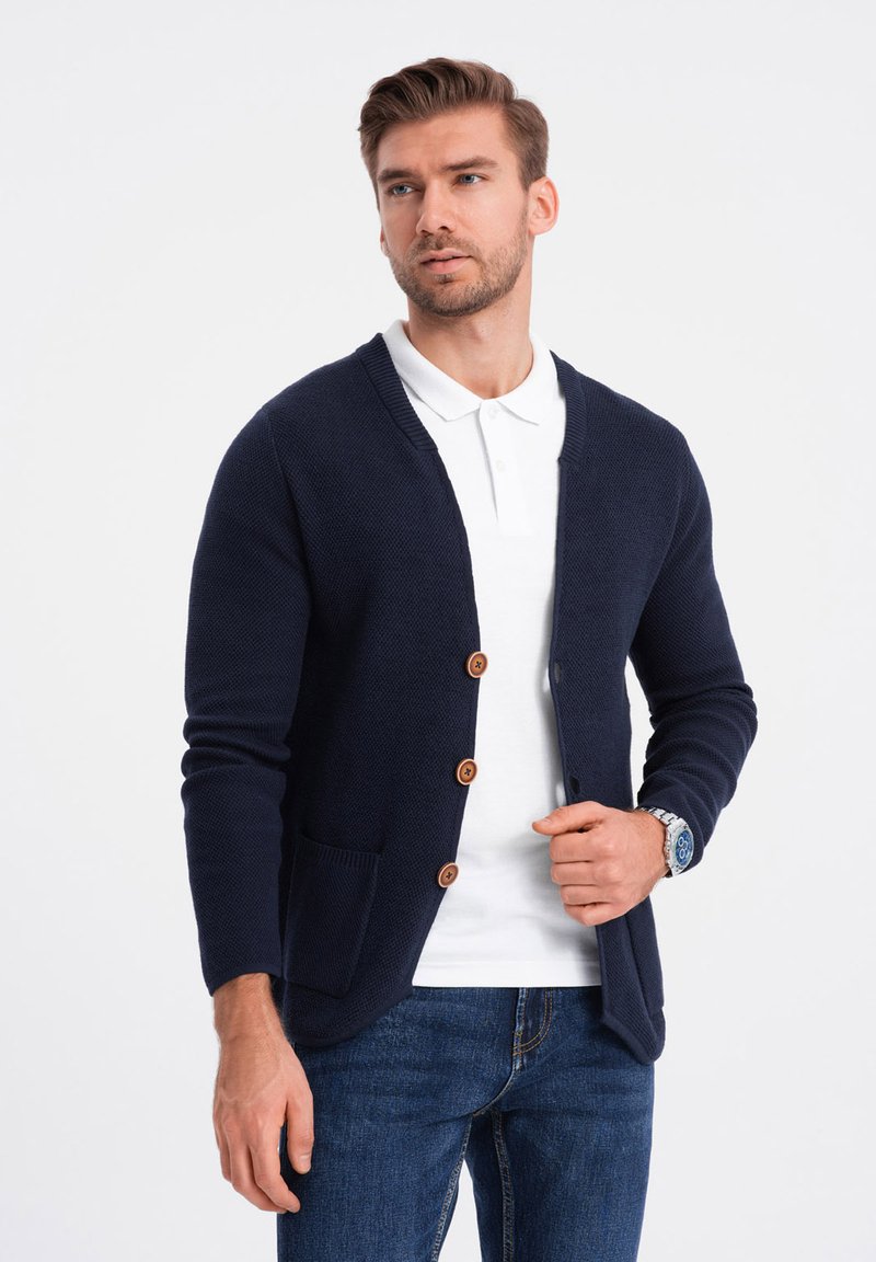 Cárdigan texturizado azul marino con botones de madera marrones, diseño en forma de v, dos bolsillos frontales, usado sobre una camiseta polo blanca y jeans de denim.