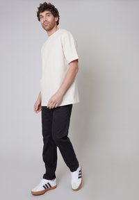 T-shirt beige a maniche corte, vestibilità rilassata, abbinato a pantaloni scuri e sneakers bianche con tre strisce nere e suole marroni.