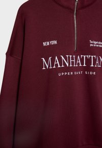 Sweatshirt bordeaux avec fermeture éclair à l'avant et texte brodé en blanc indiquant "NEW YORK", "MANHATTAN", "UPPER EAST SIDE" ainsi qu'une citation partielle.