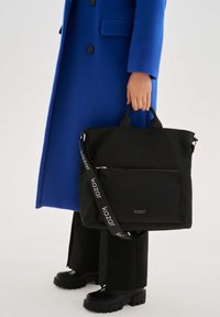 Bolsa tote de tela negra con dos asas y una correa de hombro ajustable con un logotipo. Textura suave, bolsillo con cierre en la parte frontal.