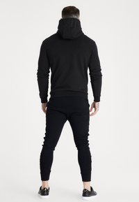 Felpa nera con cappuccio e scritte in rilievo sul colletto, abbinata a pantaloni jogger neri. Vestibilità slim, polsini a costine e cuciture dettagliate. Scarpe da ginnastica nere.