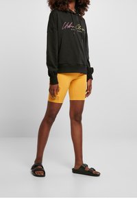 Černá mikina s kapucí s grafickým textem, spárovaná se žlutými cyklistickými šortkami a černými sandály. Má uvolněný střih a měkkou texturu.