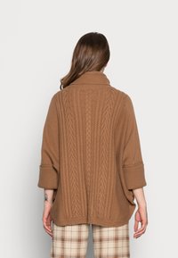 Femme aux longs cheveux bruns portant un pull ample en maille torsadée marron et un pantalon beige à carreaux, vue de dos sur un fond uni.