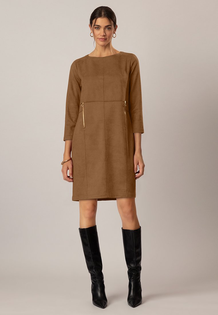 Femme portant une robe longue à manches longues de couleur brune, arrivant au genou, avec des détails de zip dorés et des bottes noires jusqu'au genou, sur un fond uni.