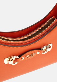 Pomarańczowa torebka z teksturowanego materiału syntetycznego, z akcentami w kolorze złotym i zapięciem na zamek. Posiada dekoracyjną łańcuszek oraz detal z logo.