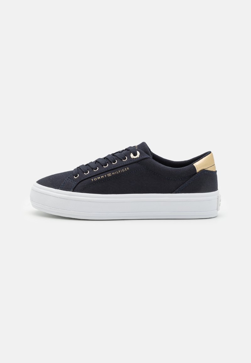 Navy canvas sneakers with white rubber sole, gold accent on heel, and gold eyelets. Feature flat laces and textured fabric detail.