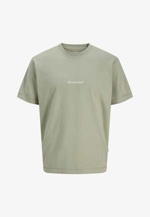 Garçon adolescent debout, portant un t-shirt vert clair avec un texte blanc, un pantalon noir et des baskets beige et blanches, sur un fond clair uni.