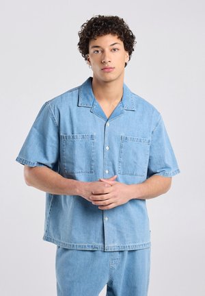 Jonge man met krullend haar, gekleed in een lichtblauw denim shirt met korte mouwen en bijpassende broek, staand met samengevouwen handen.