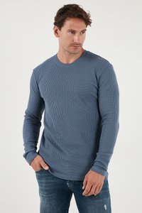 Uomo che indossa una camicia blu a maniche lunghe con trama e jeans blu, in piedi con una mano in tasca, che guarda verso destra.