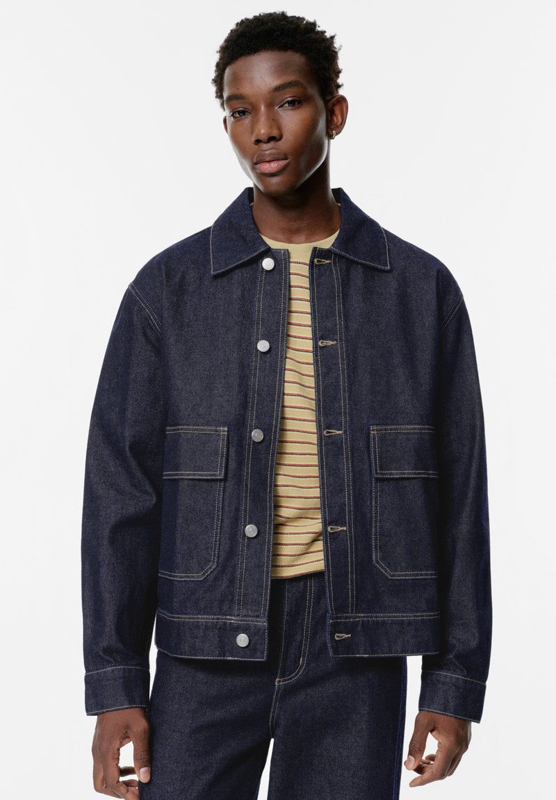 Giacca in denim scuro con due tasche frontali, bottoni argentati e colletto, indossata sopra una camicia a righe chiara. Texture liscia e vestibilità su misura.