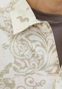 Camisa con patrón floral que presenta intrincados diseños en beige y rosa pálido sobre una tela suave, con un cuello estándar y detalle de botones en el cuello.