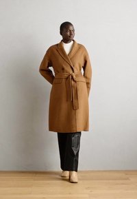 Brauner Wollmantel mit einem Taillengürtel, doppelreihiger Knopfleiste und seitlichen Taschen, kombiniert mit schwarzen Hosen und beigen Stiefeletten.