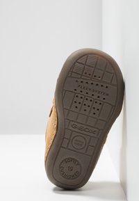 Scarpa in suede marrone con suola in gomma flessibile e imbottita. La suola presenta fori di ventilazione e un marchio in rilievo. Sono visibili dettagli cuciti.