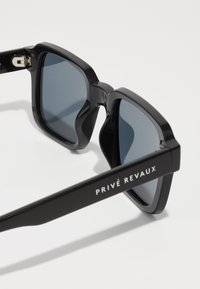 Occhiali da sole neri con una montatura audace e angolare, lenti scure e una finitura lucida. Presentano il nome del marchio "PRIVÉ REVAUX" sul braccio.
