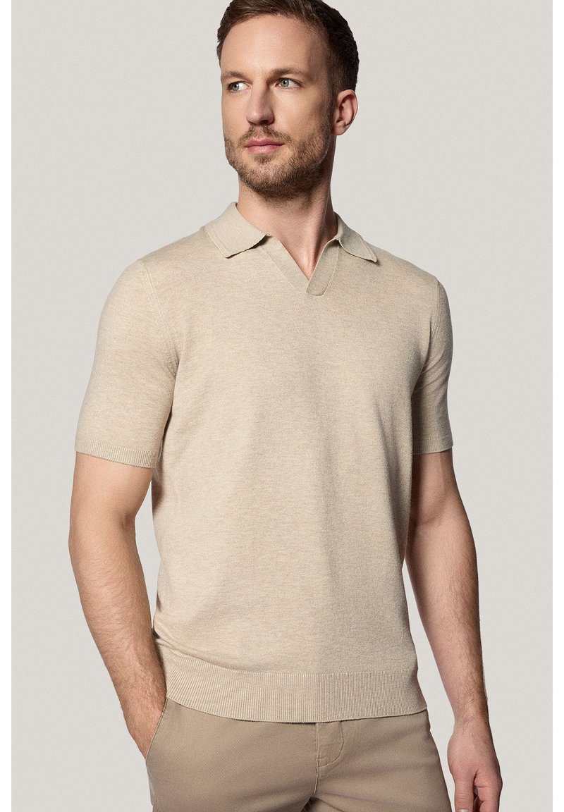 Beżowa, krótko rękawowa koszulka polo z dekoltem w kształcie V, z prążkowanymi mankietami i dołem, wykonana z miękkiego, teksturowanego materiału, noszona z neutralnymi spodniami.