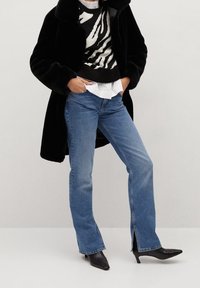 Osoba v černém kabátu z umělé kožešiny, s pulovrem se zebrovým vzorem přes bílou košili, modrými džíny s rozparkem a černými špičatými lodičkami s nízkým podpatkem.