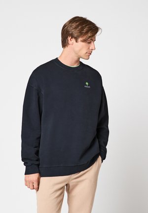 Homme portant un sweat-shirt noir à col rond avec un petit logo d'arbre vert et un pantalon beige, regardant vers le bas à sa gauche.