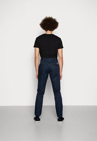 Jeune personne aux cheveux bouclés se tient face à un mur blanc, portant un t-shirt noir, un pantalon bleu foncé et des baskets Converse noires.