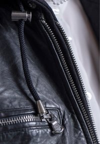 Zwarte leren jas met een gestructureerde afwerking, ritssluitingen en metalen accenten. Inclusief een zwarte trekkoord dat aan de capuchon vastzit.