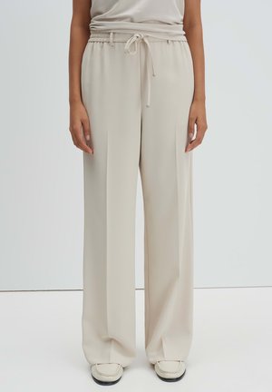 Person trägt lockere, beigefarbene Hosen mit Kordelzug und Falten, kombiniert mit weißen Slip-on-Schuhen vor einfarbigem Hintergrund.