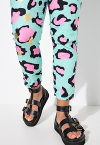 Turquoise pants with a black and pink leopard print, featuring a slightly crinkled texture. Black strappy sandals with buckles and a thick sole.