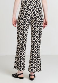 Personne portant un pantalon géométrique noir et beige vu de dos, associé à des ballerines noires à talon ouvert.