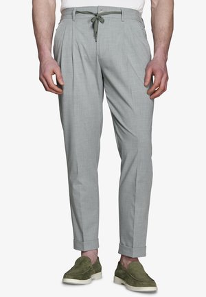 Mężczyzna w jasno szarych, dopasowanych spodniach z elastycznym paskiem i zielonych zamszowych butach wsuwanych, stojący na białym tle.