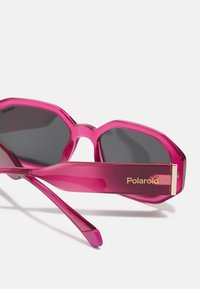 Gafas de sol hexagonales rosas con lentes oscuros. Montura de plástico con textura brillante. Marca "Polaroid" en dorado en la patilla.