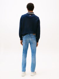 Göndör hajú férfi, aki tengerészkék pulóvert, világoskék farmert és fehér tornacipőt visel, háttal áll egy fehér háttér előtt.