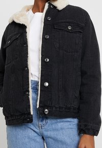 Svart denimjacka med krämfärgad fleecefoder, tryckknappar och dubbla bröstfickor, med avslappnad passform och medellång design.