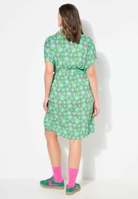 Abito verde con motivo floreale circolare. Tessuto morbido, maniche corte, vita con cintura. Calzini rosa accesi e scarpe verdi con dettagli rosa.