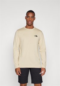 Beige langermet t-skjorte laget av bomull, med en svart logo på venstre bryst, kombinert med svarte shorts. Enkelt design, standard passform.