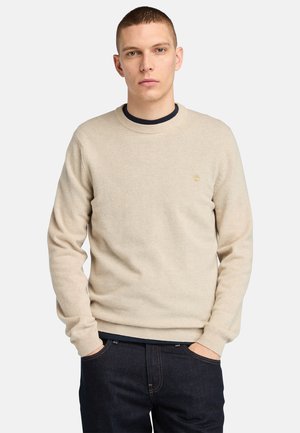 Maglione beige con scollatura rotonda e polsini a coste. Presenta un piccolo logo sul petto. Abbinato a jeans scuri. Texture morbida.