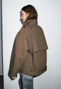 Brown oversized jacket with a high collar, shoulder epaulets, back flap detail, and side pockets, paired with light textured jeans.