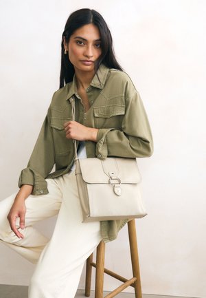 Bēša ādas soma ar vāciņu un sudraba detaļām, ko rokās tur persona, kura valkā olīvu zaļas krāsas krekls un krēmkrāsas bikses, sēžot uz krēsla.