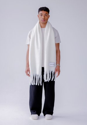 Witte gebreide sjaal met franjes aan de uiteinden en een zwart verloopmotief. Het label draagt de tekst "ARCTIC FOX." Draagt over een lichtgrijs shirt.