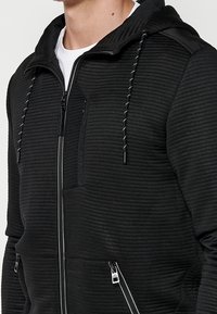 Felpa nera con zip, con motivo a coste e trama strutturata, cappuccio regolabile con coulisse e tasche con cerniera. Presenta dettagli a strisce e logo.