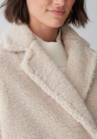 Beige Fleecejacke mit strukturierter Oberfläche, ausgestattet mit einem breiten Kragen und einem offenen Design, getragen über einem hellcremefarbenen Pullover.